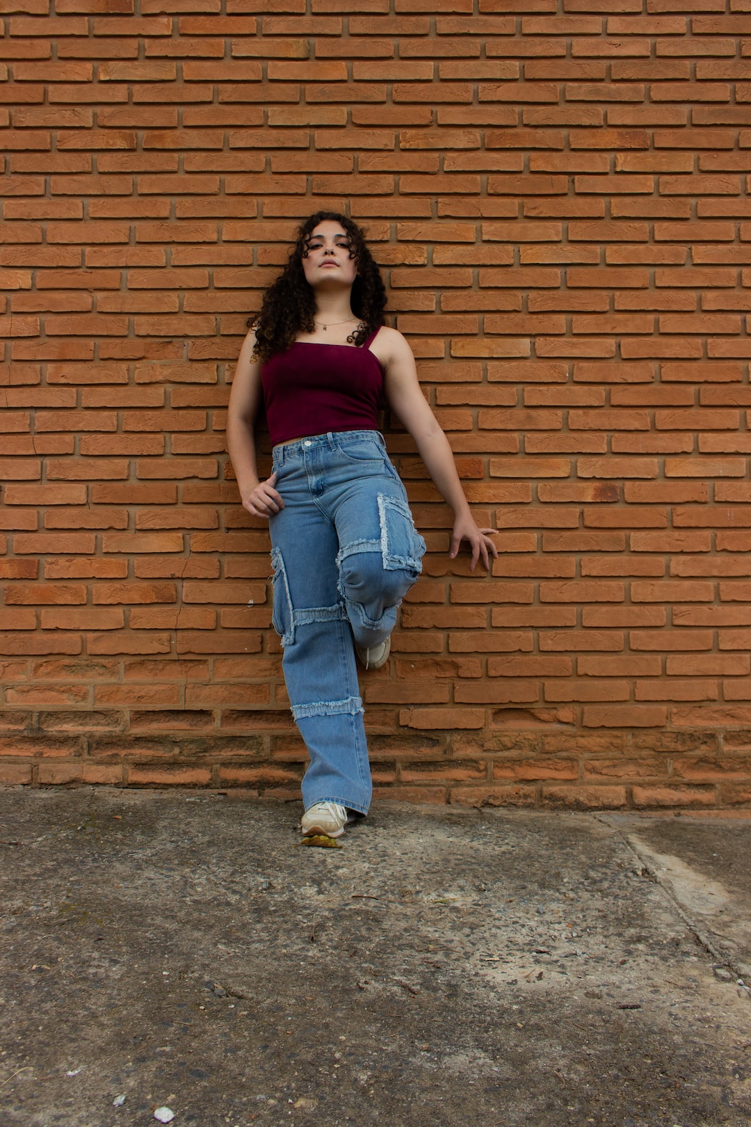 woman in red tank top and blue denim jeans standing beside brown brick wall
