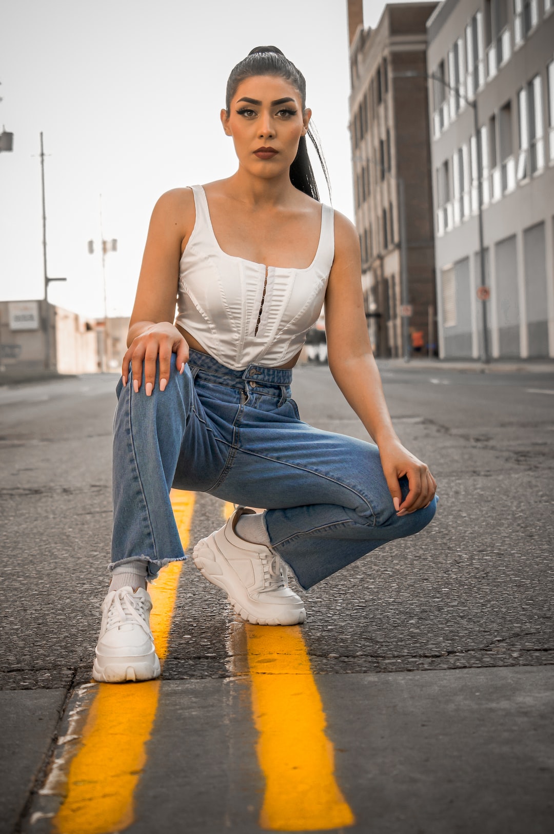 woman in white tank top and blue denim jeans sitting on gray concrete road during daytime
